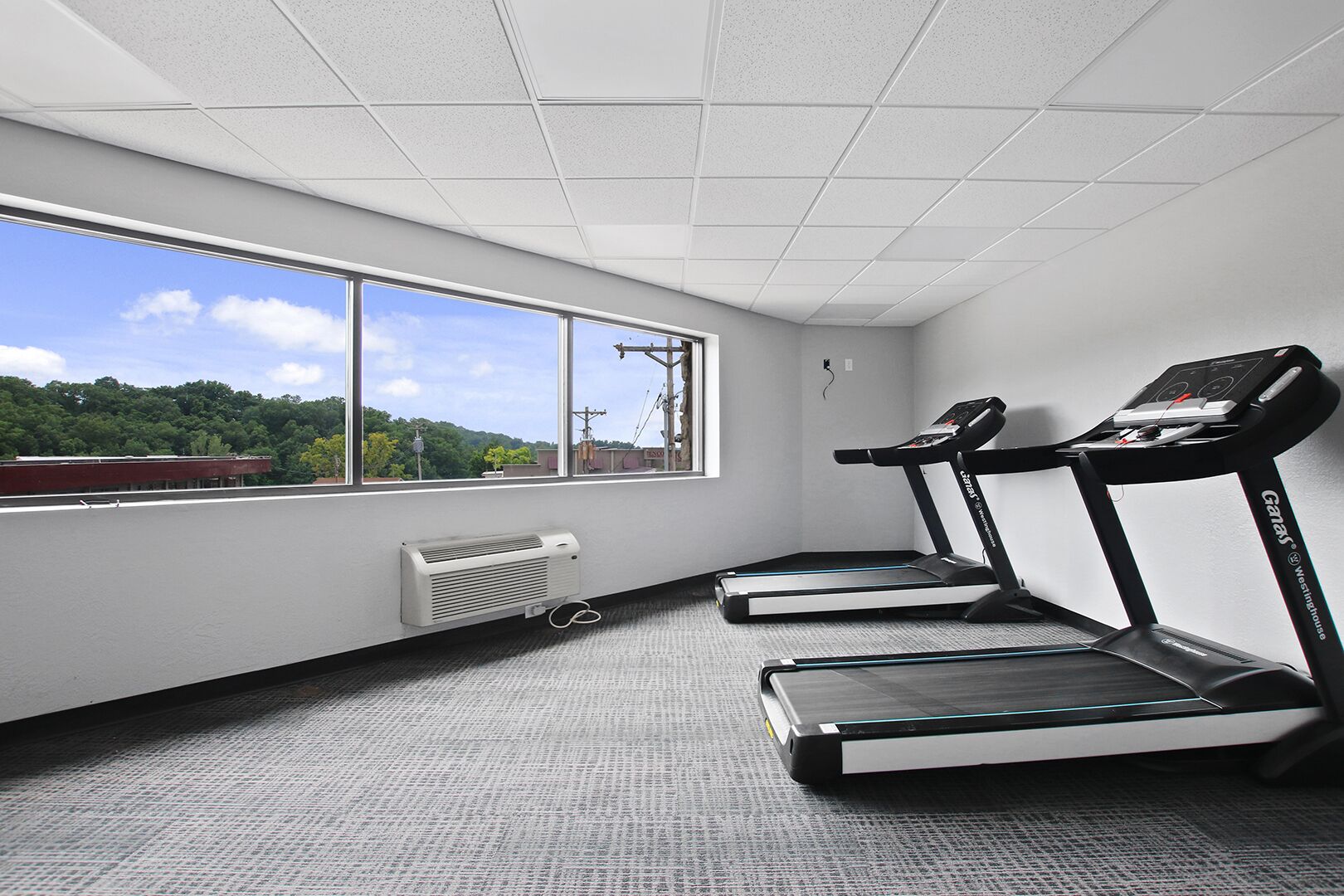 A gym with two treadmills, a window with a view of trees and buildings, and a ceiling with a grid pattern.