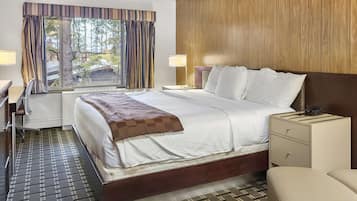 A hotel room with a large bed, a desk, and a chair. There is a window with curtains, a nightstand, and a lamp.