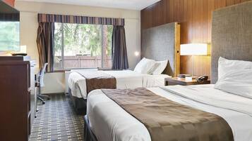 A hotel room with two beds, a desk, a chair, a lamp, and a view of greenery outside.