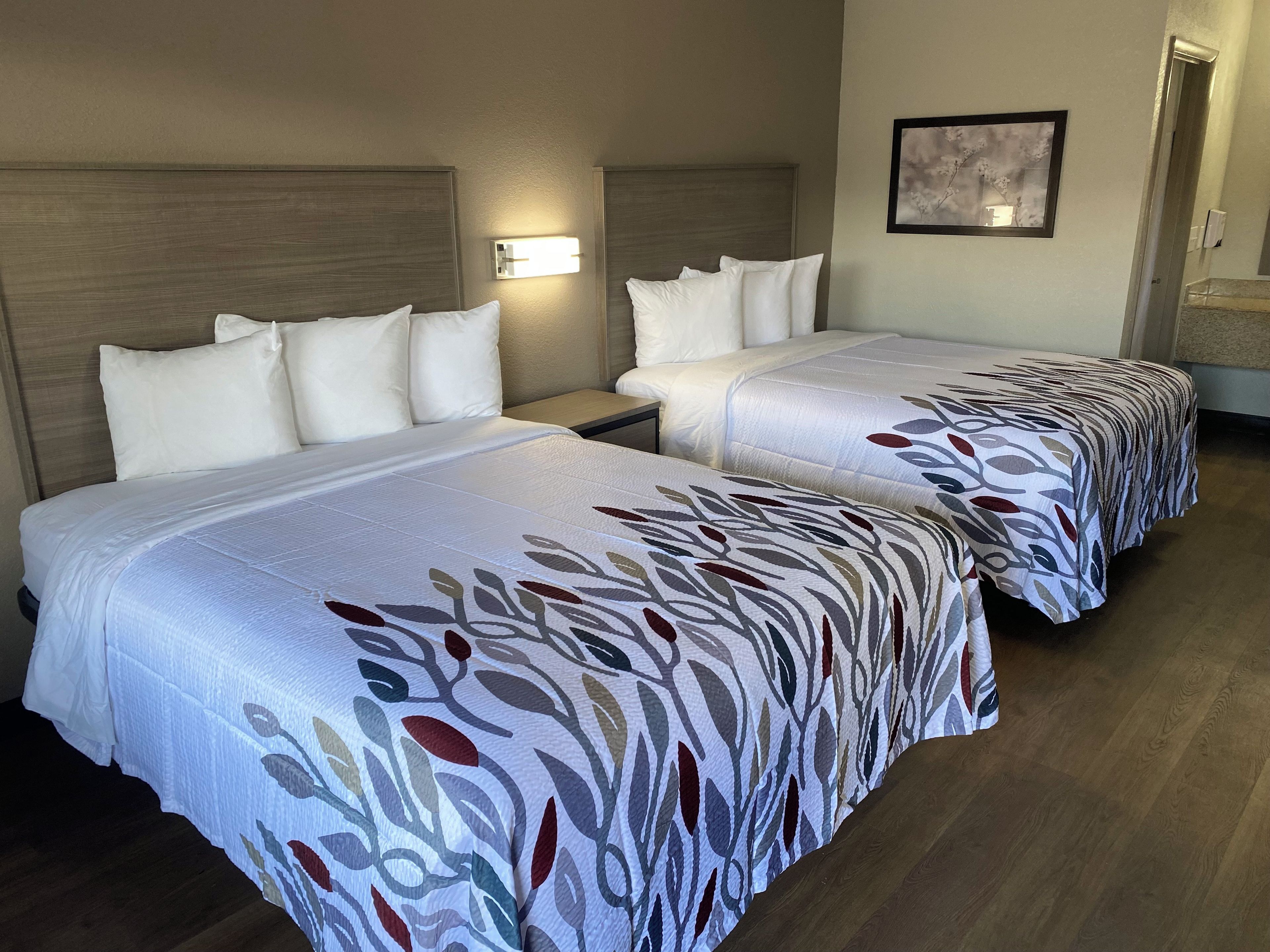 A hotel room with two beds, each with a patterned bedspread, a nightstand, a wall-mounted light, and a framed picture on the wall.