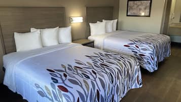 A hotel room with two beds, each with a patterned bedspread, a nightstand, a wall-mounted light, and a framed picture on the wall.