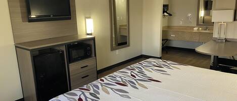 A hotel room with a bed, a TV mounted on the wall, a desk with a microwave, and a bathroom visible through an open door.