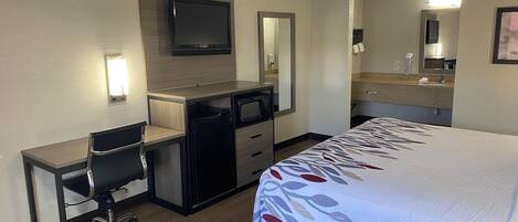 Ein Hotelzimmer mit einem Bett, einem Schreibtisch, einem Sessel, einem Fernseher, einer Mikrowelle und einem Badezimmer mit Waschbecken und Spiegel.