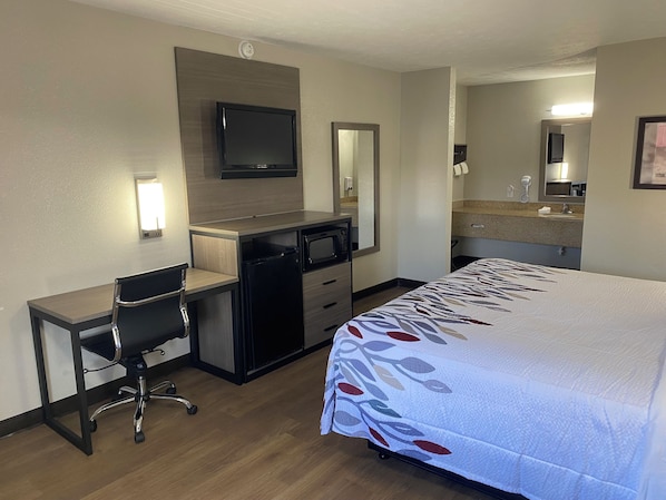 Quarto de hotel com cama, escrivaninha, cadeira, televisão, micro-ondas e banheiro com pia e espelho.