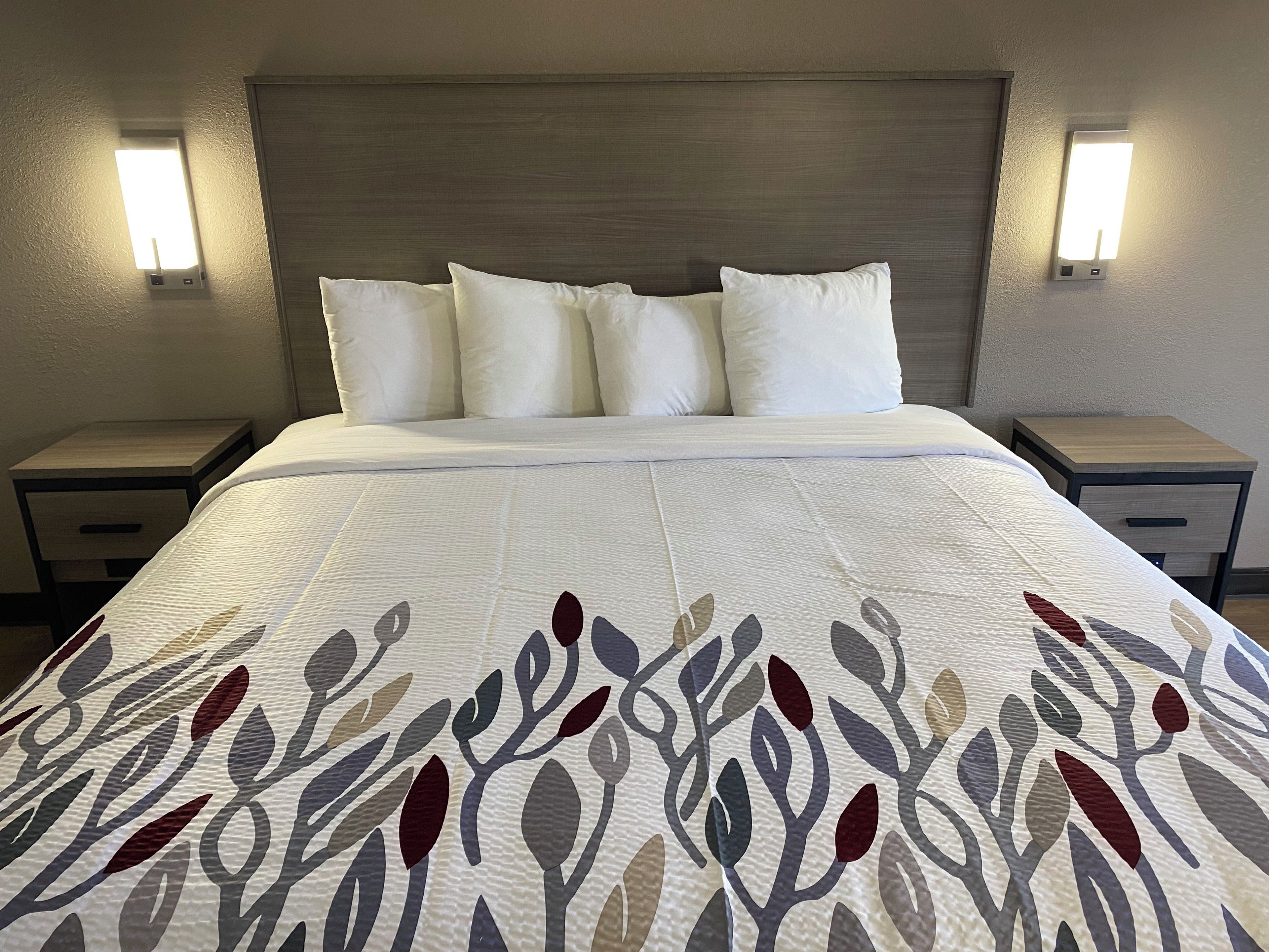 Habitación de hotel con cama, ropa blanca de cama, colcha estampada, dos mesitas de noche e iluminación mural.