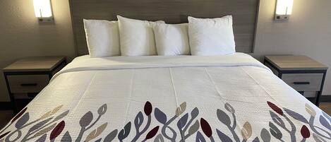 Habitación de hotel con cama, ropa blanca de cama, colcha estampada, dos mesitas de noche e iluminación mural.