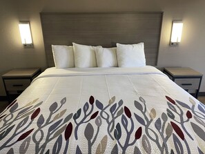 A hotel room with a bed, white bedding, a patterned bedspread, two bedside tables, and wall-mounted lights. - Red Roof Inn Jacksonville, AL  (Jacksonville)