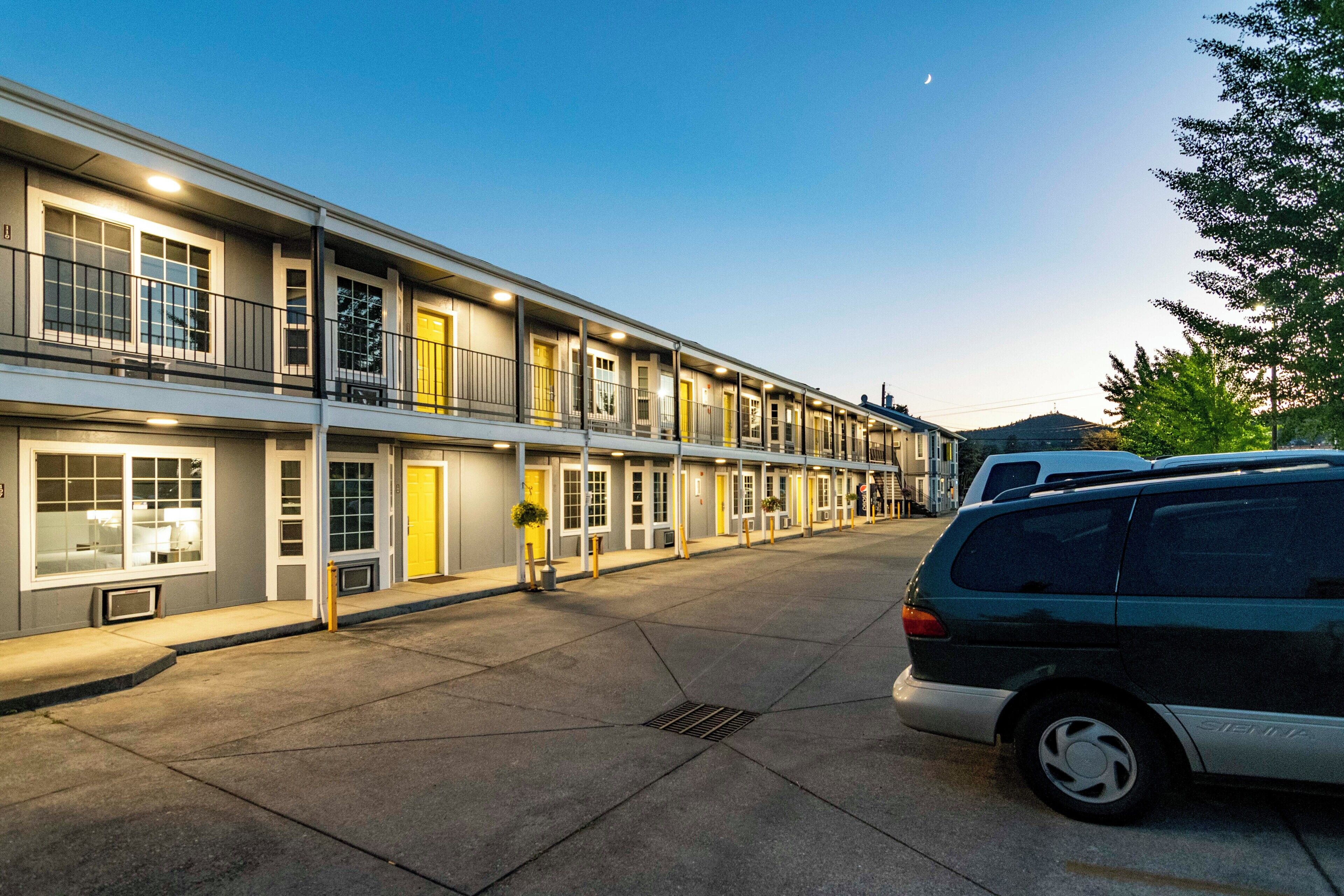 Un hotel de varios pisos con una fila de habitaciones, un estacionamiento y una furgoneta estacionada al frente.