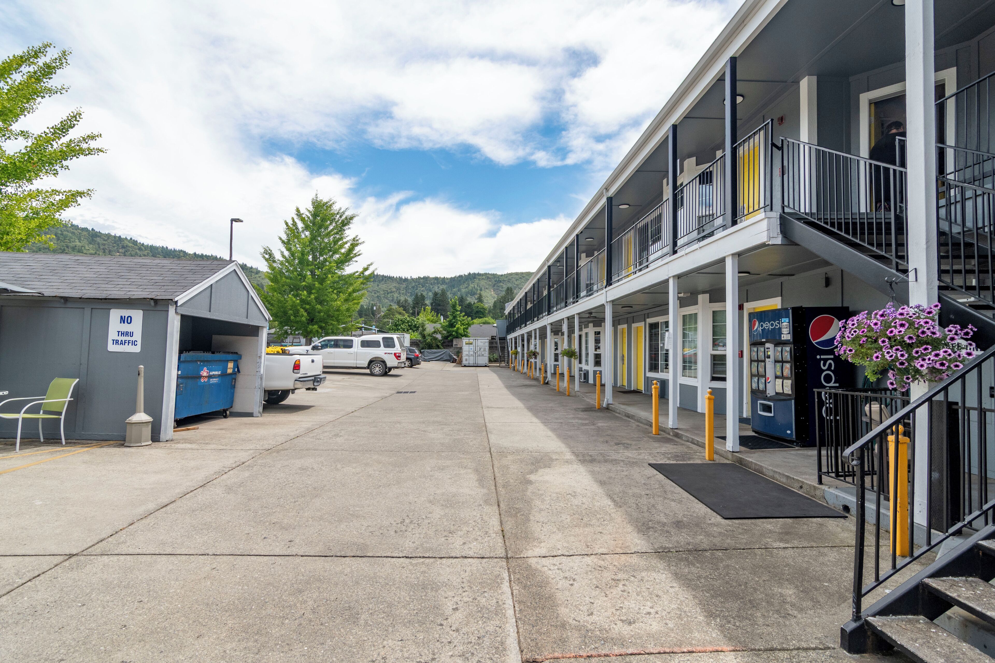 Um motel com estacionamento, uma lata de lixo, uma máquina de venda automática de Pepsi e uma escadaria que dá acesso ao andar superior.