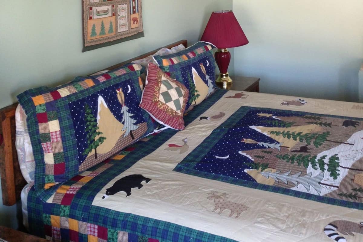 A bedroom with a bed, a quilt, a wall hanging, a lamp, and a shelf.