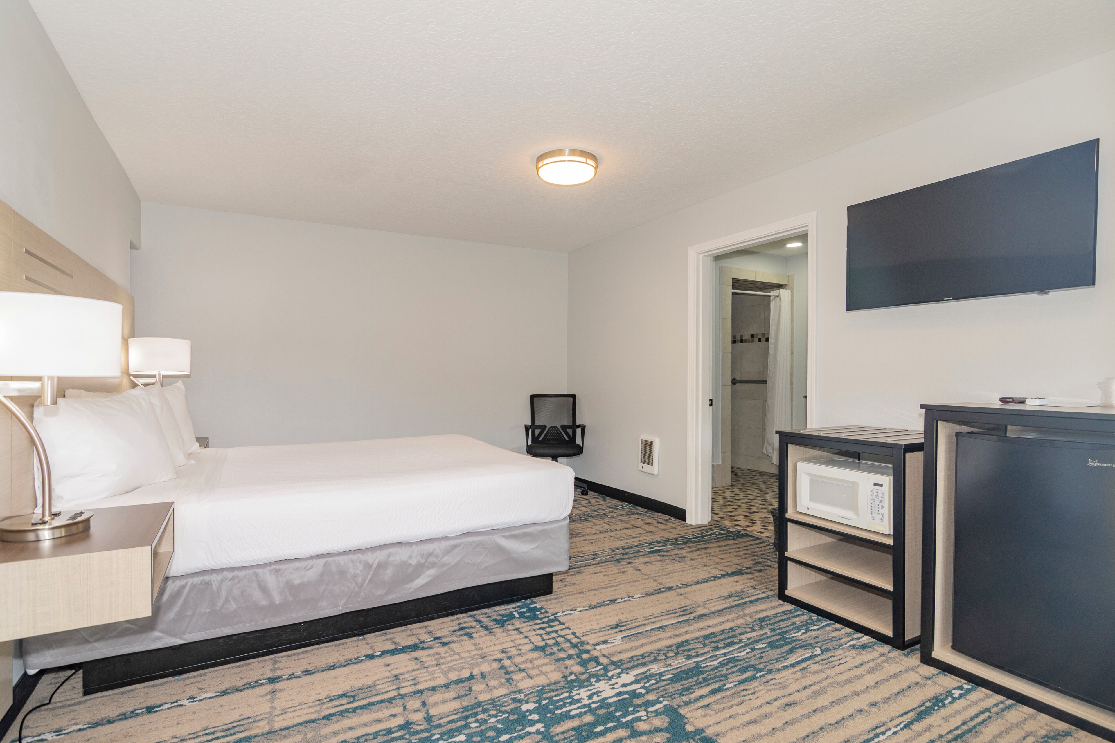 A hotel room with a bed, bedside table, lamp, TV, microwave, and a door leading to a bathroom.