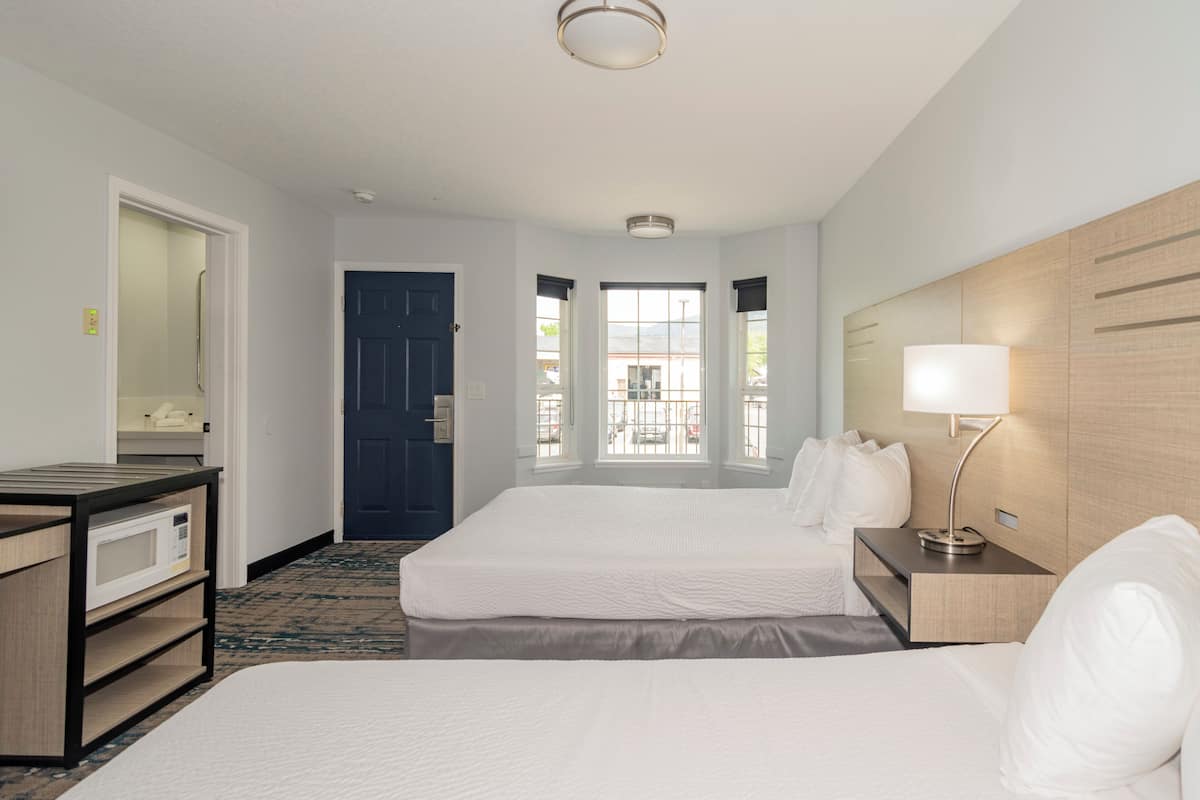 A hotel room with two beds, a desk, a lamp, a microwave, and a view of the outside through large windows.