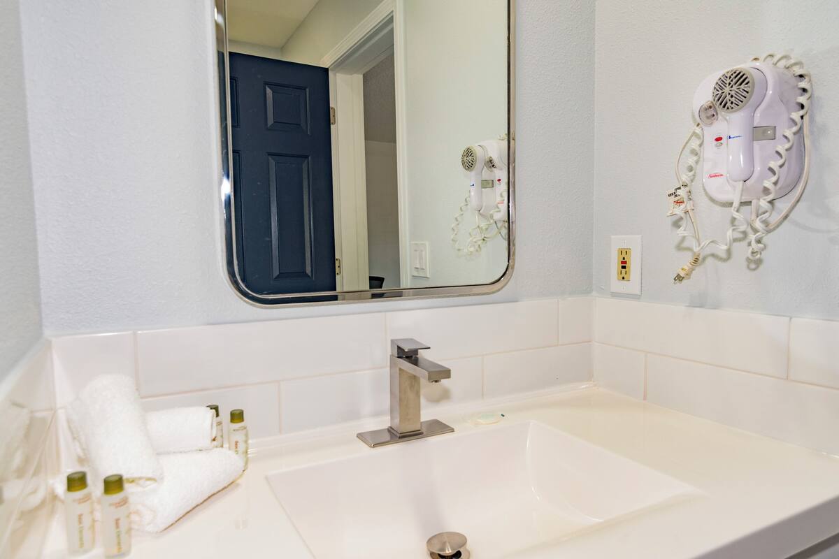 A bathroom with a white sink, a mirror, a hair dryer mounted on the wall, and a door leading to another room.