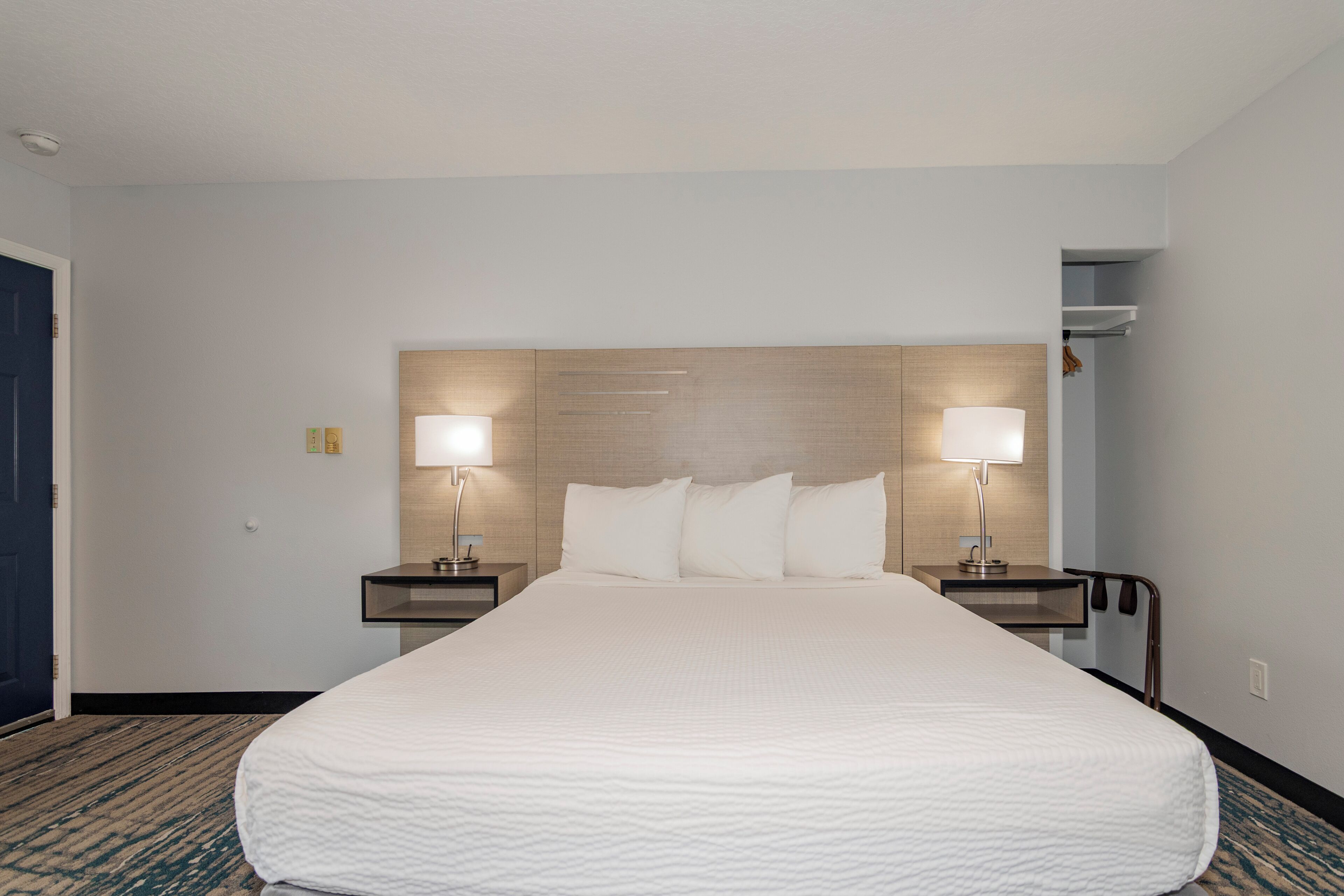 A hotel room with a large bed, two bedside lamps, a wooden headboard, and a carpeted floor.