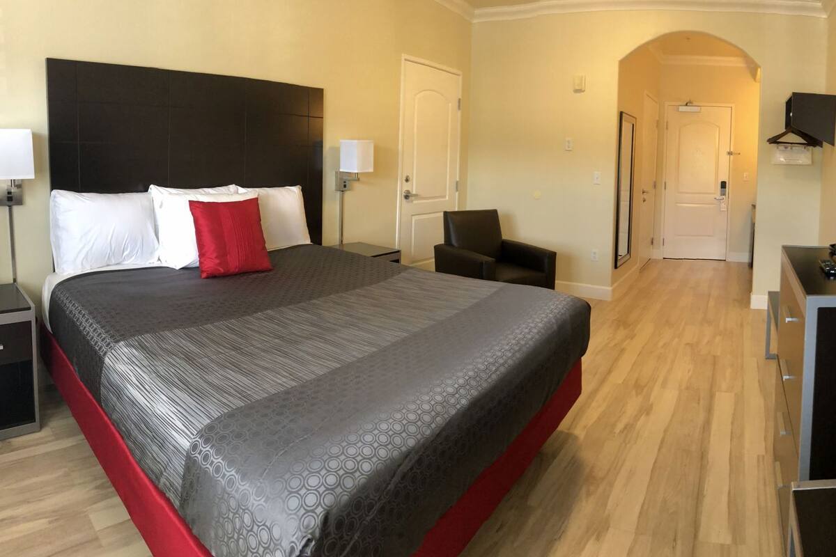 A hotel room with a large bed, bedside tables, a desk, a chair, a TV, and a door leading to another room.