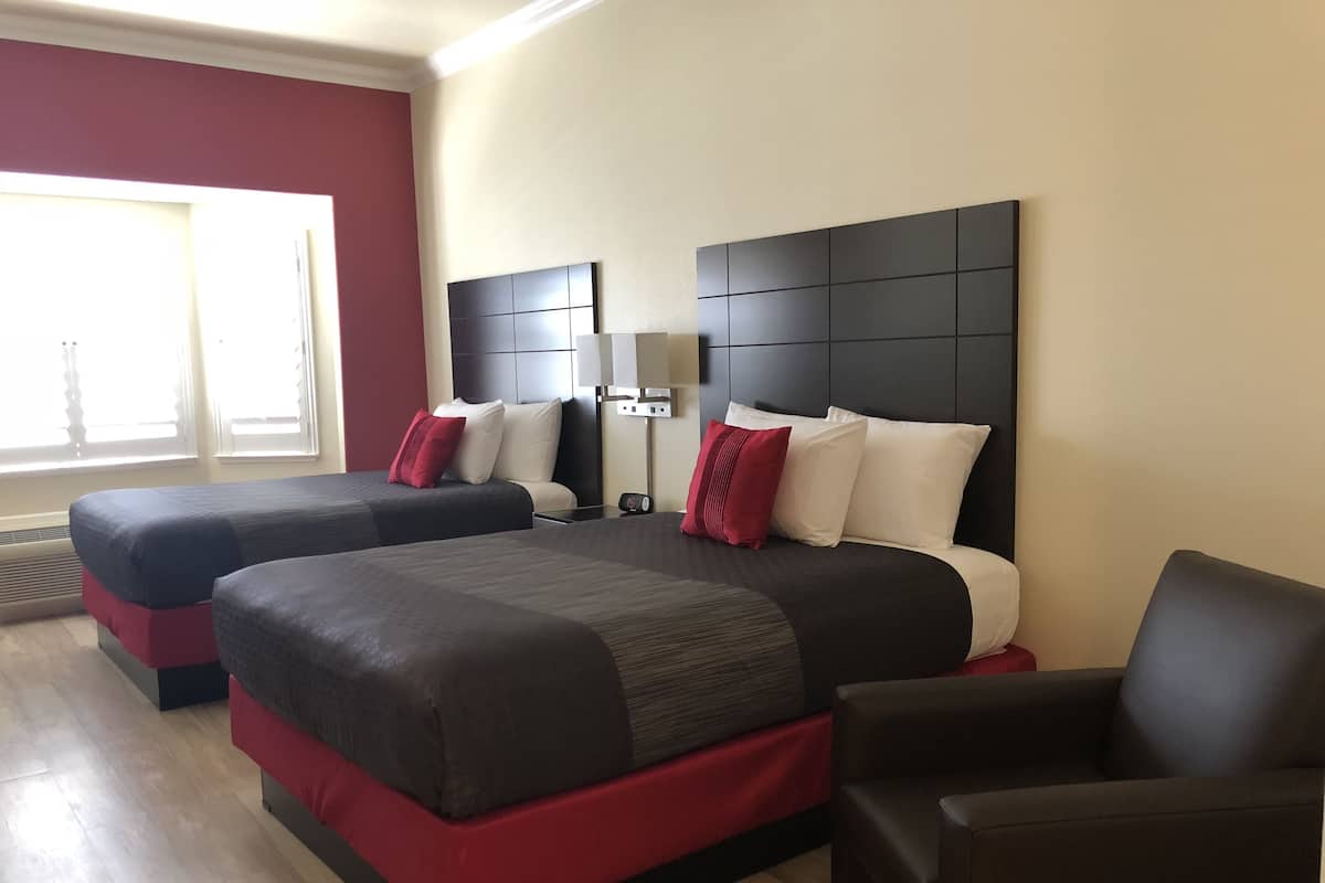 A hotel room with two beds, a red accent wall, a large window, and a modern design.