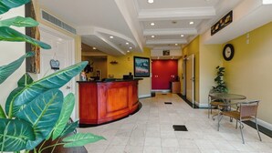 A hotel lobby with a reception desk, a dining area with wicker chairs, and a red wall with a clock. - Gateway Inn & Suites San Francisco SFO Airport (San Bruno)