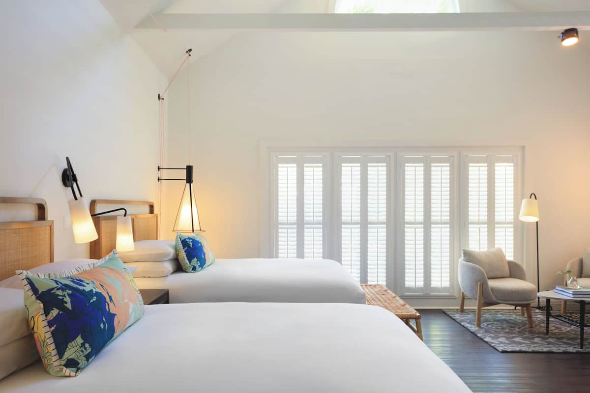 A modern hotel room with two beds, a sitting area, and a large window with blinds.