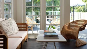 A room with a wooden sofa, a wicker chair, a small white table, and a view of a balcony with outdoor furniture.