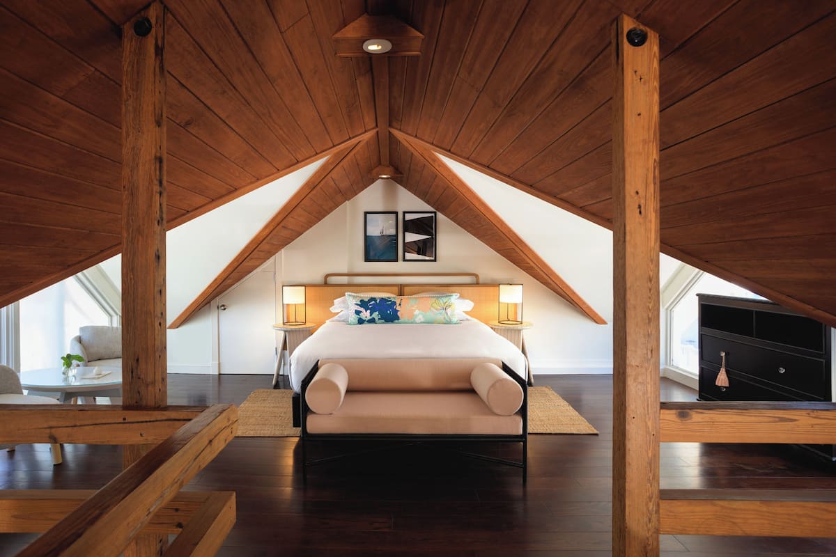 A bedroom with a sloped ceiling, wooden beams, a bed with a white headboard, two bedside lamps, and a large window.