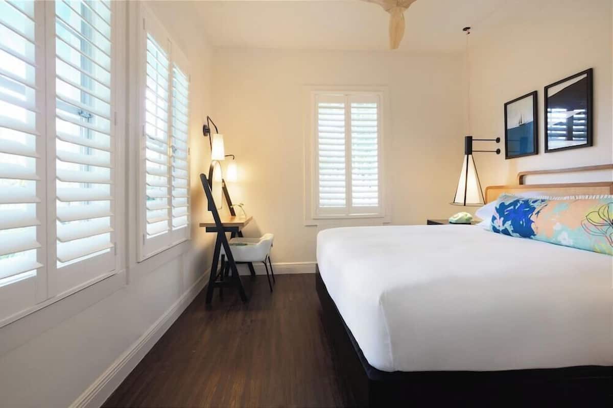 A bedroom with a bed, a desk, a chair, and two windows with shutters.