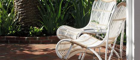 Een witte rieten stoel met metalen frame, geplaatst op een metselstenen terras met een witte hek en groene begroeiing op de achtergrond.