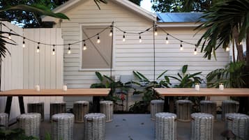 An outdoor patio with two wooden tables and matching stools, surrounded by plants and string lights.