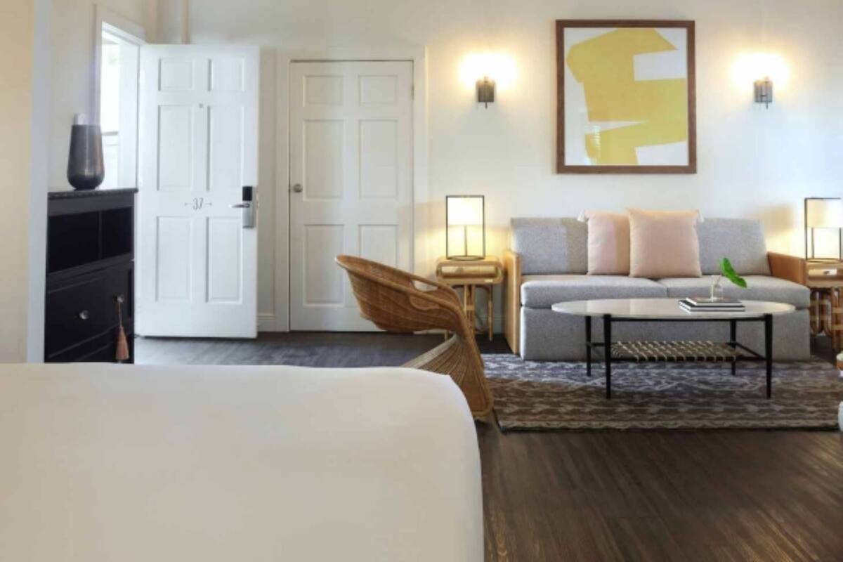 A spacious room with a large bed, a sofa, a chair, and a desk. There are two wall-mounted lamps and a framed abstract painting.