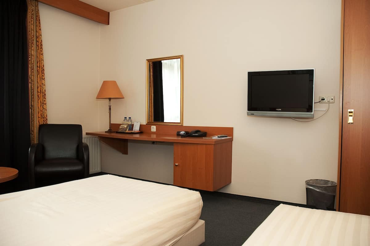 A hotel room with a bed, a desk with a lamp, a chair, a TV, and a trash can.