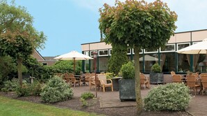 An outdoor seating area with wicker chairs and tables, surrounded by greenery and a building with large windows. - Fletcher Hotel Steenwijk (Steenwijk)