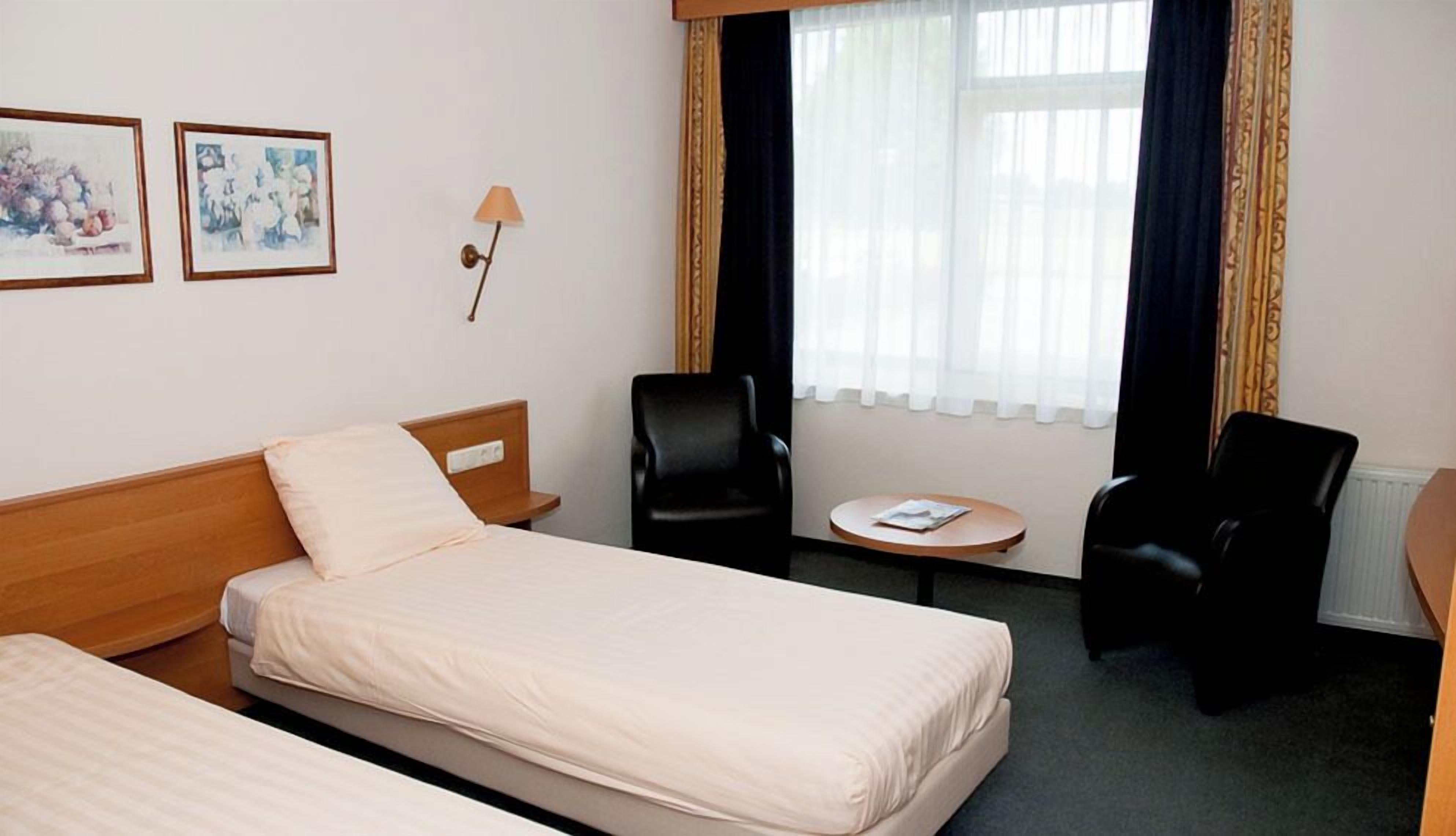 A hotel room with two beds, a small table, two chairs, a lamp, and two framed pictures on the wall.