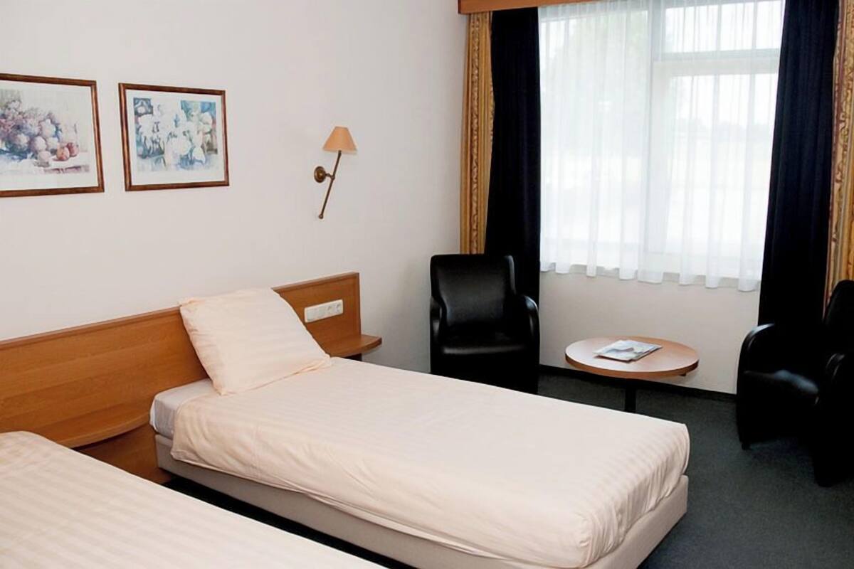 A hotel room with two beds, a small table, two chairs, a lamp, and two framed pictures on the wall.