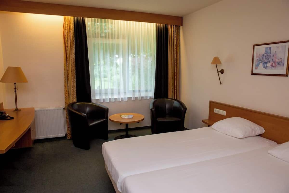 A hotel room with a bed, two chairs, a small table, a lamp, a radiator, and a window with curtains.