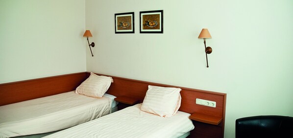 A hotel room with two single beds, a wooden headboard, two wall-mounted lamps, and two framed pictures on the wall.