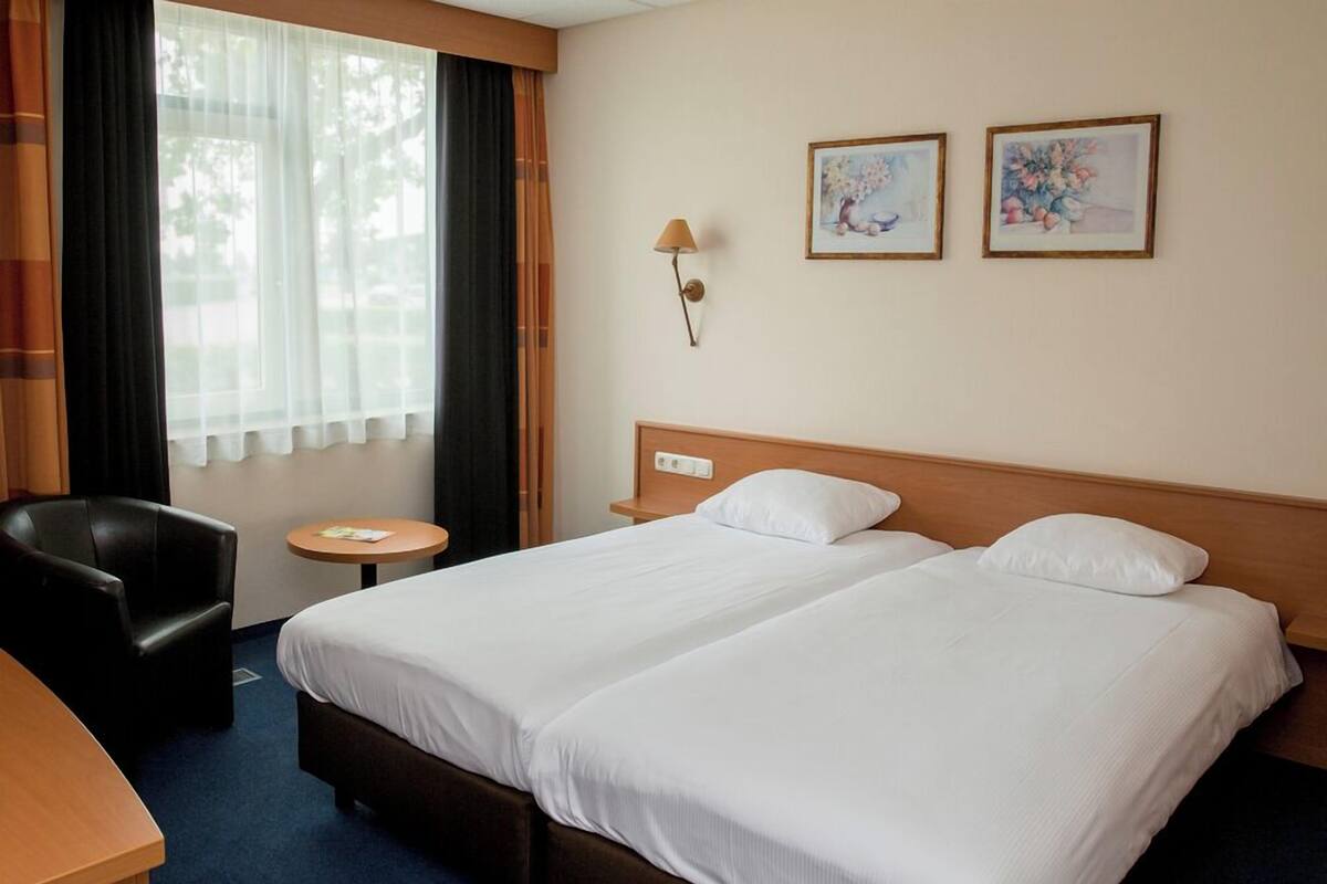 A hotel room with two beds, a desk, a chair, a small table, and a window with curtains.