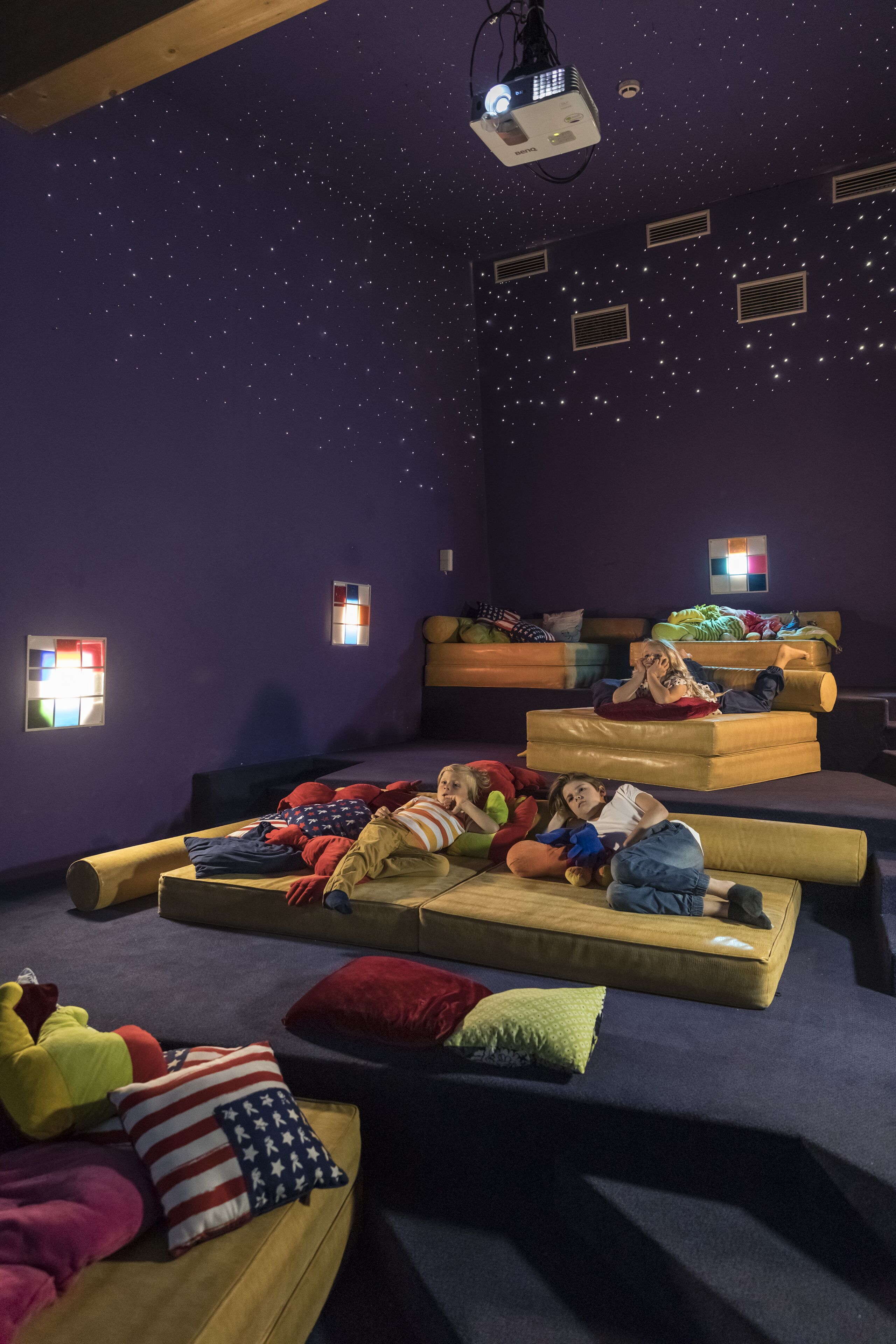 A room with a starry ceiling, multiple beds, and small windows with colorful light panels.
