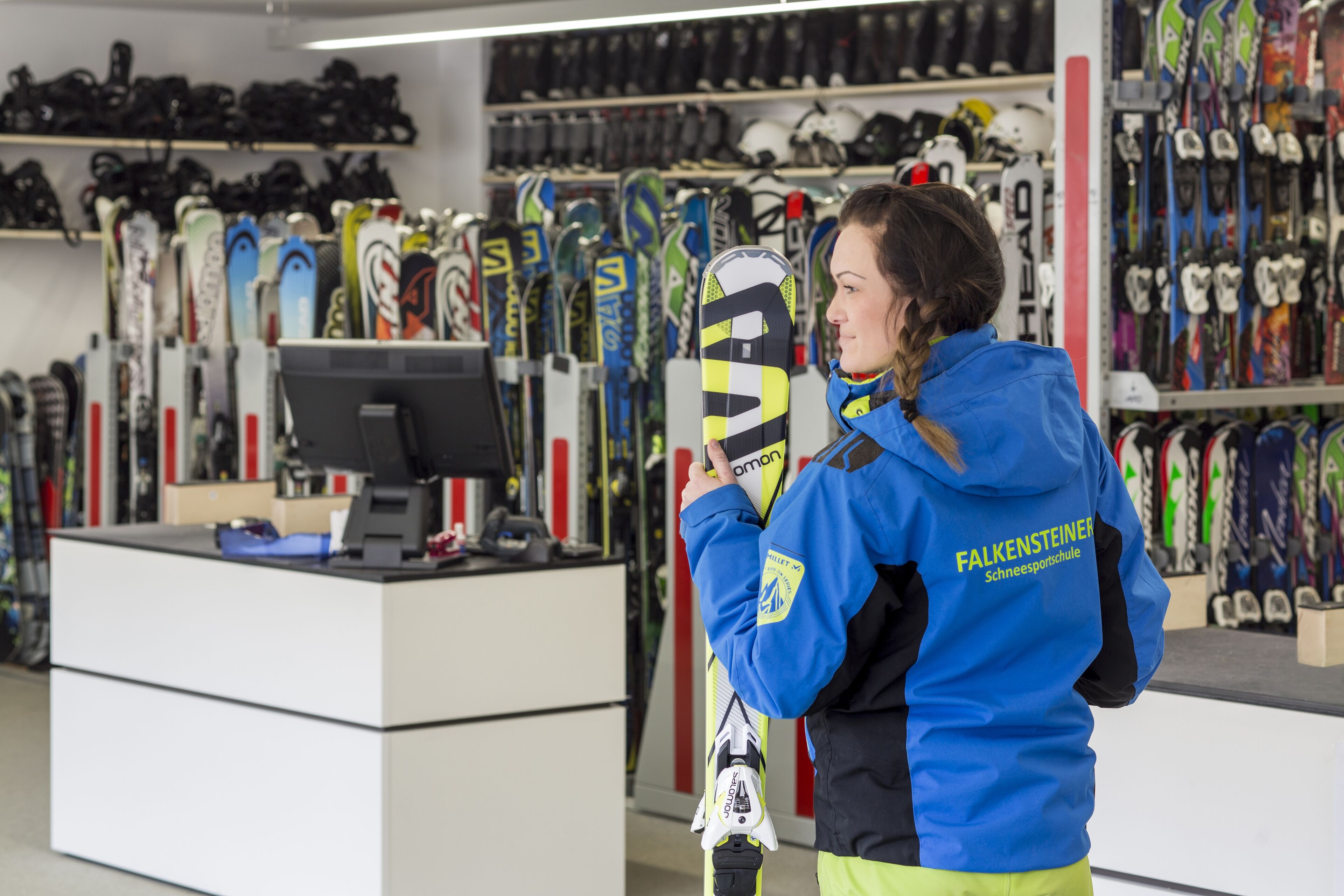 A person in a blue jacket holding skis in a ski shop with various skis on display.