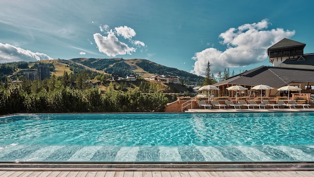 A swimming pool with a wooden deck, lounge chairs, and umbrellas, overlooking a mountainous landscape with buildings. - Falkensteiner Club Funimation Katschberg (Rennweg am Katschberg)
