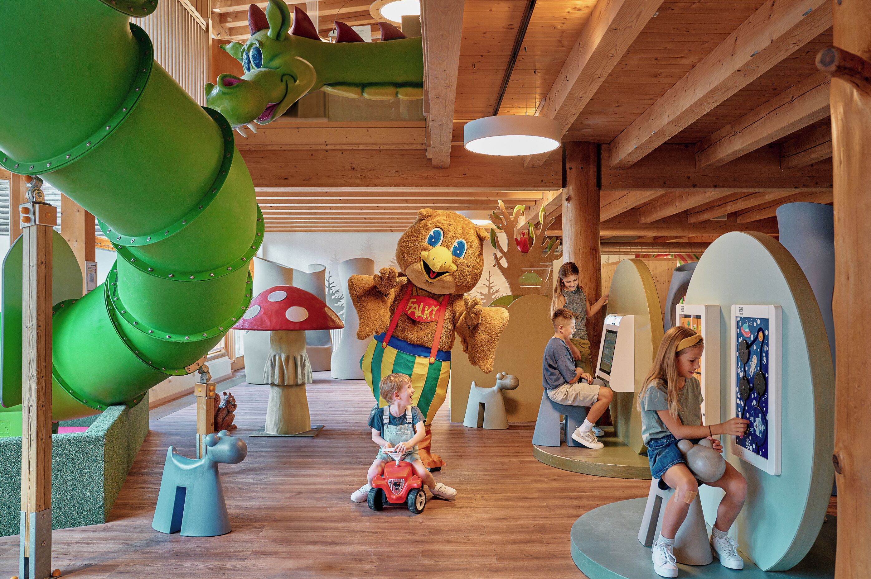 Ein Spielbereich mit einer grünen Rutsche, einem Bären als Maskottchen und spielenden Kindern.