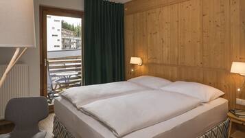 Una cama doble con cabecera de madera, una silla, una mesita de noche con lámpara, y vista a un edificio a través de una ventana con cortinas verdes.