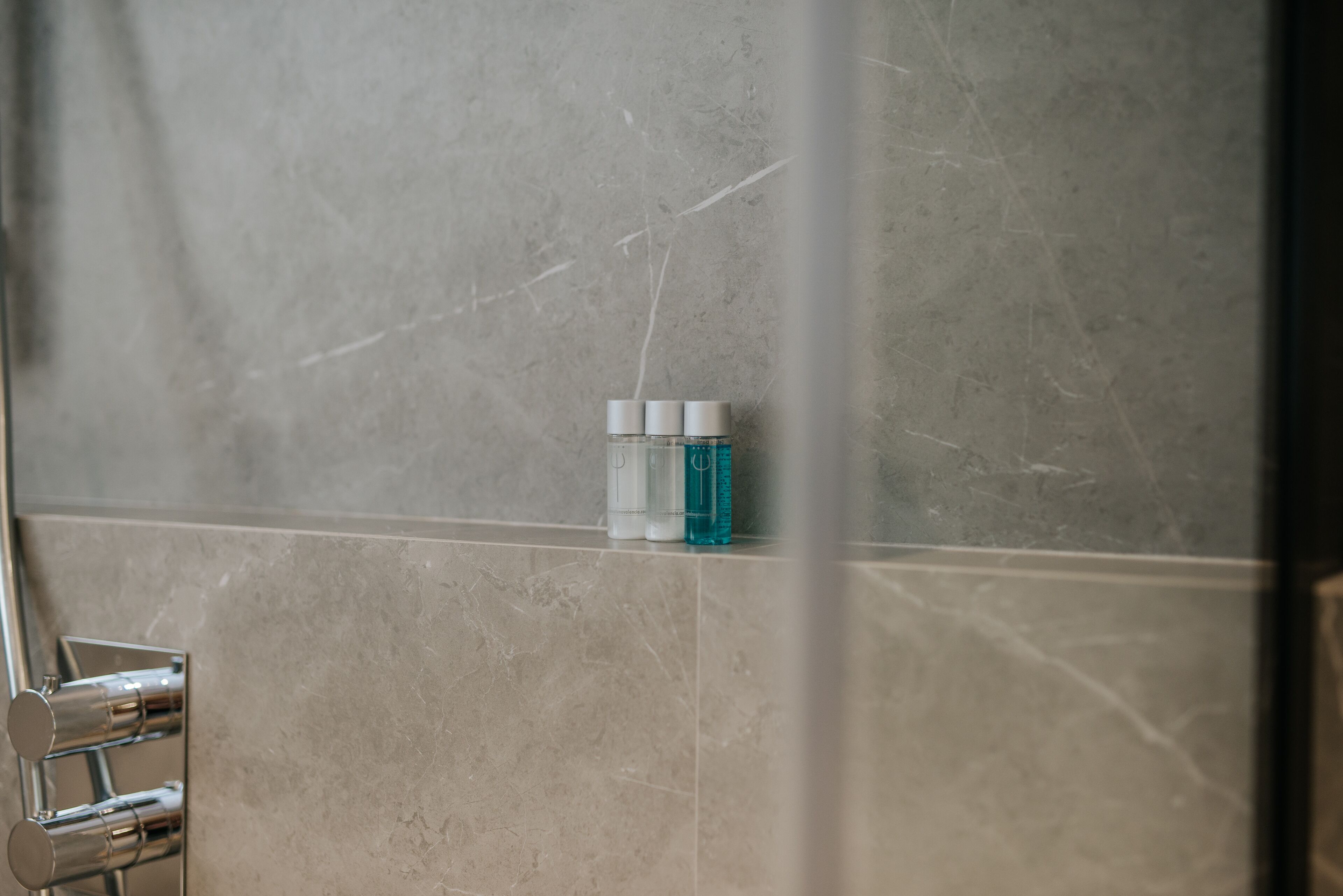 Un baño con pared de mármol, zona de ducha y estante con artículos de aseo.