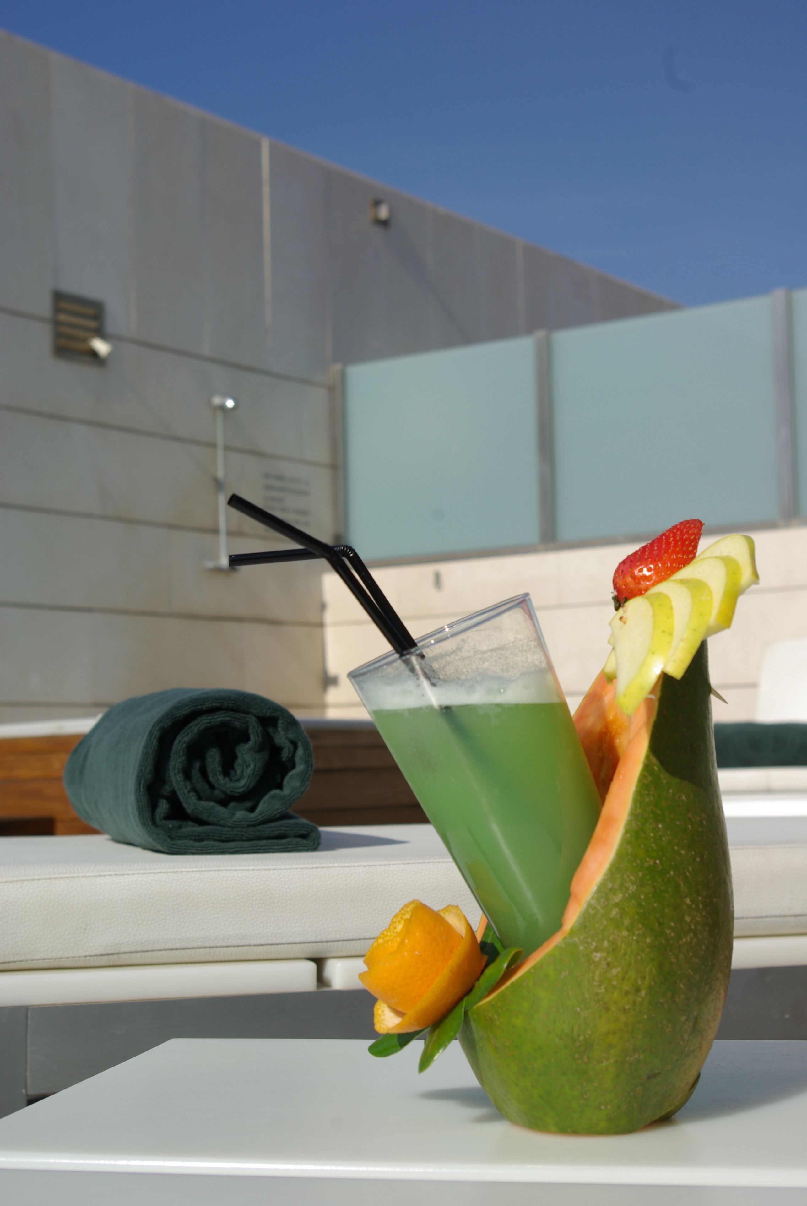 Cóctel verde en un vaso singular con decoración de frutas, una toalla enrollada y una mesa blanca frente a la fachada de un edificio moderno.