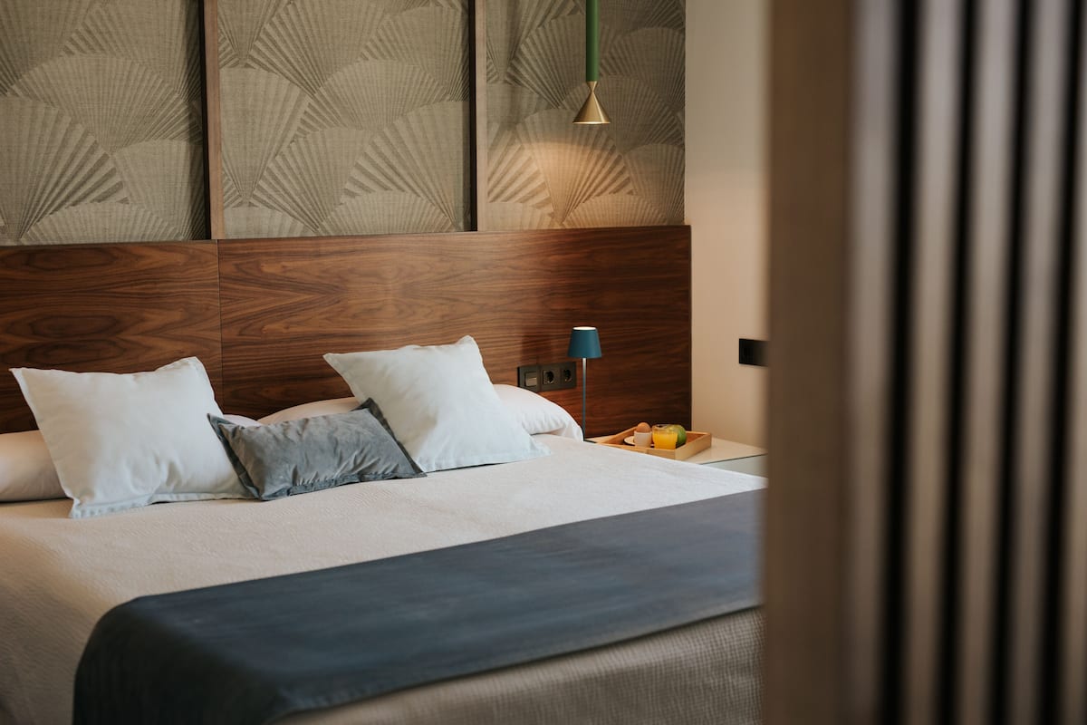 Una cama bien hecha con cabecera de madera, una mesita de noche con lámpara y una bandeja con frutas.