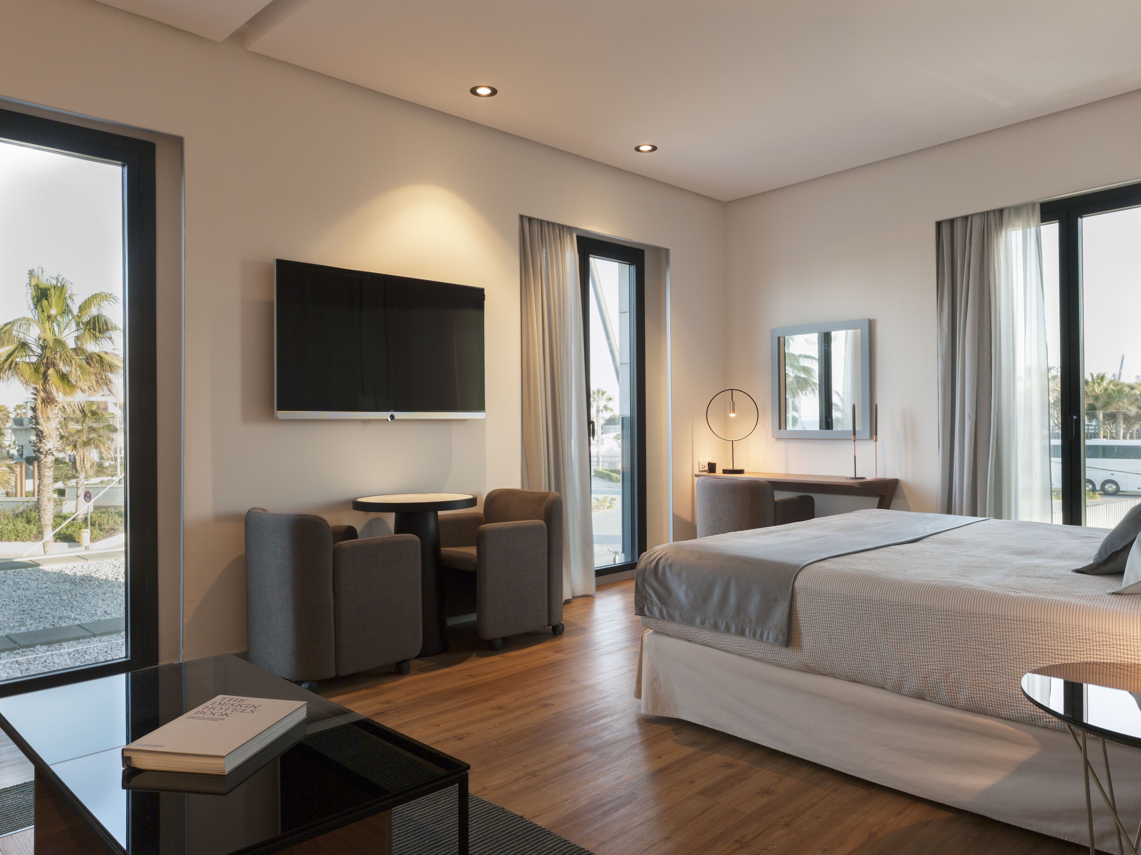 A modern hotel room with a large bed, a desk with two chairs, a television, and a large window with curtains.