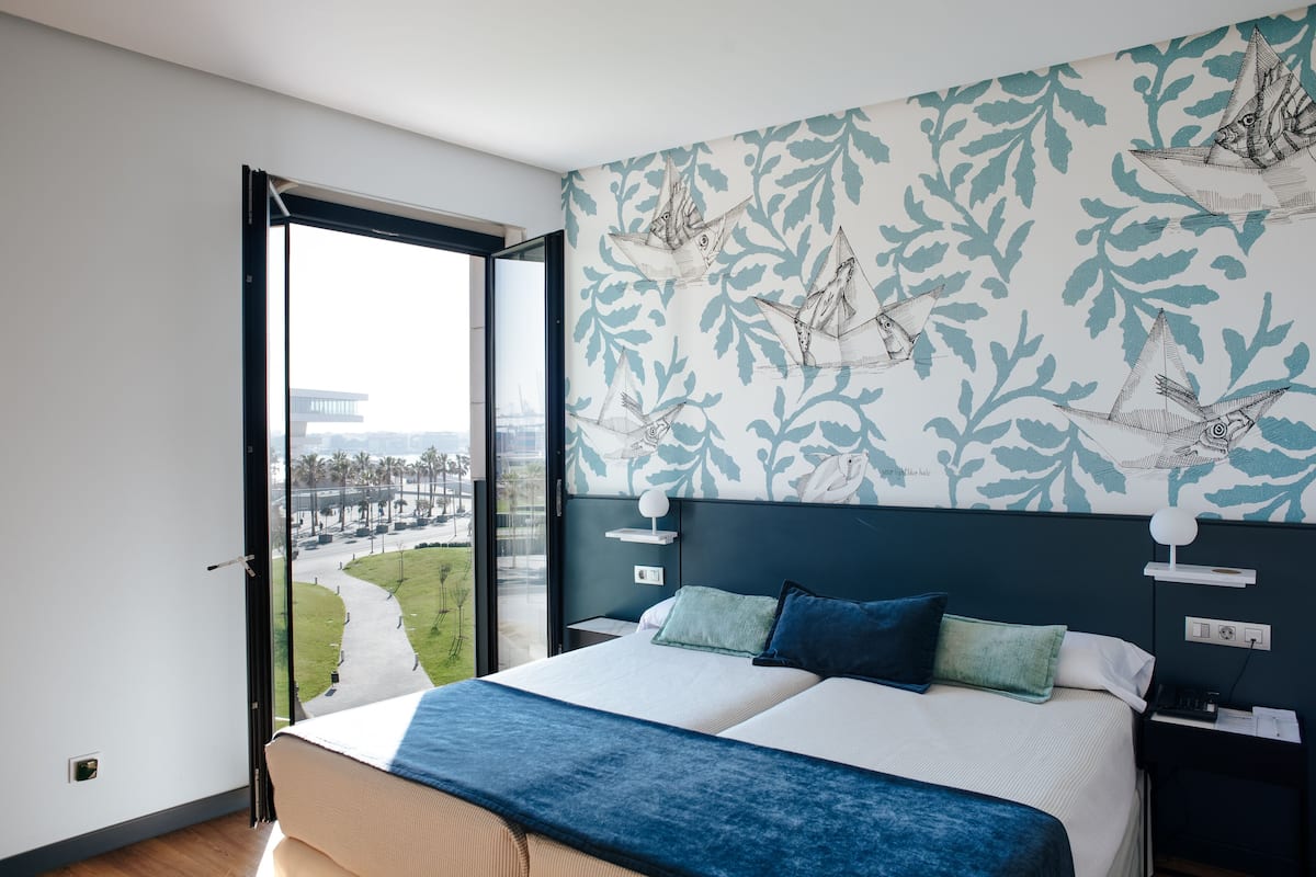 Habitación de hotel moderna con una cama grande, vista de un paisaje urbano a través de una puerta abierta y un papel tapiz estampado.