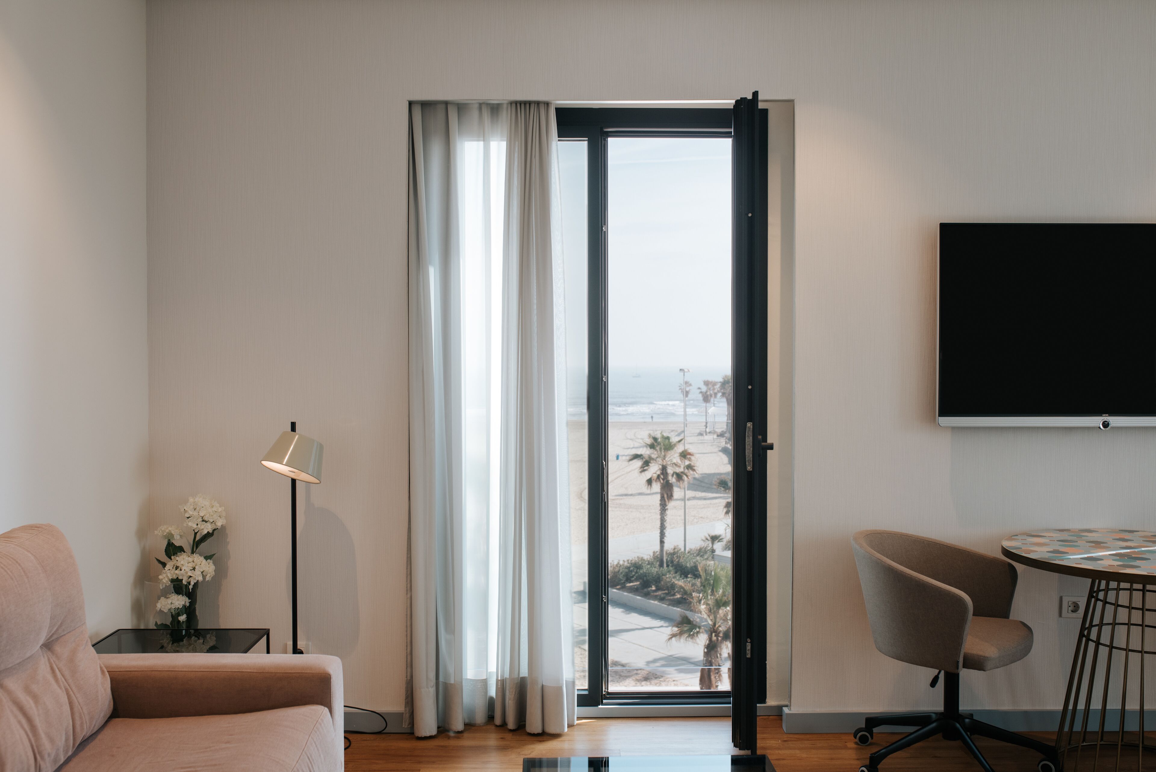 Une chambre d’hôtel moderne dotée d’une grande porte-fenêtre coulissante donnant sur un balcon, d’un téléviseur à écran plat fixé au mur, d’un bureau avec une chaise et d’un canapé.