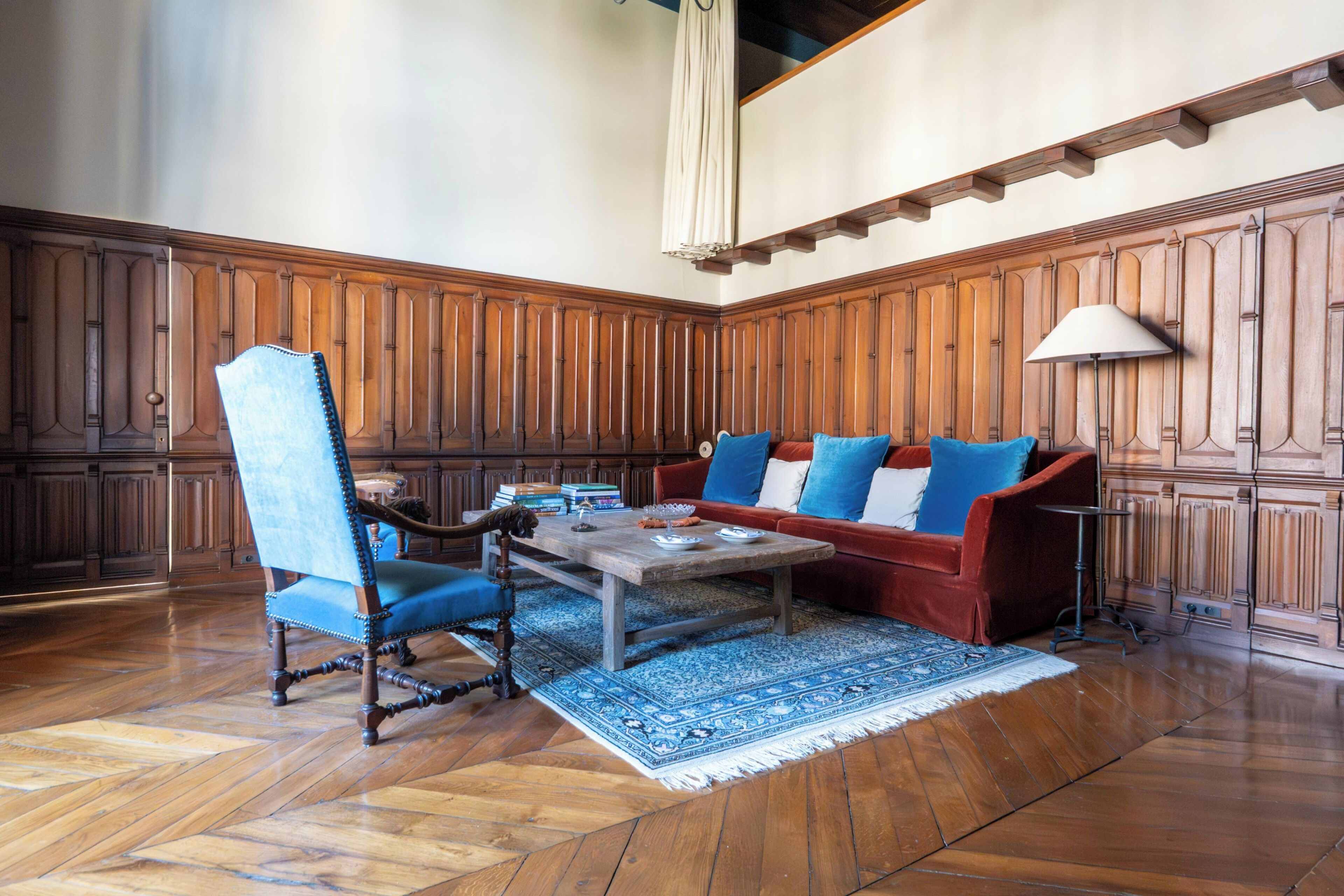 A room with a wooden paneled wall, a red sofa with blue cushions, a blue armchair, and a wooden coffee table.