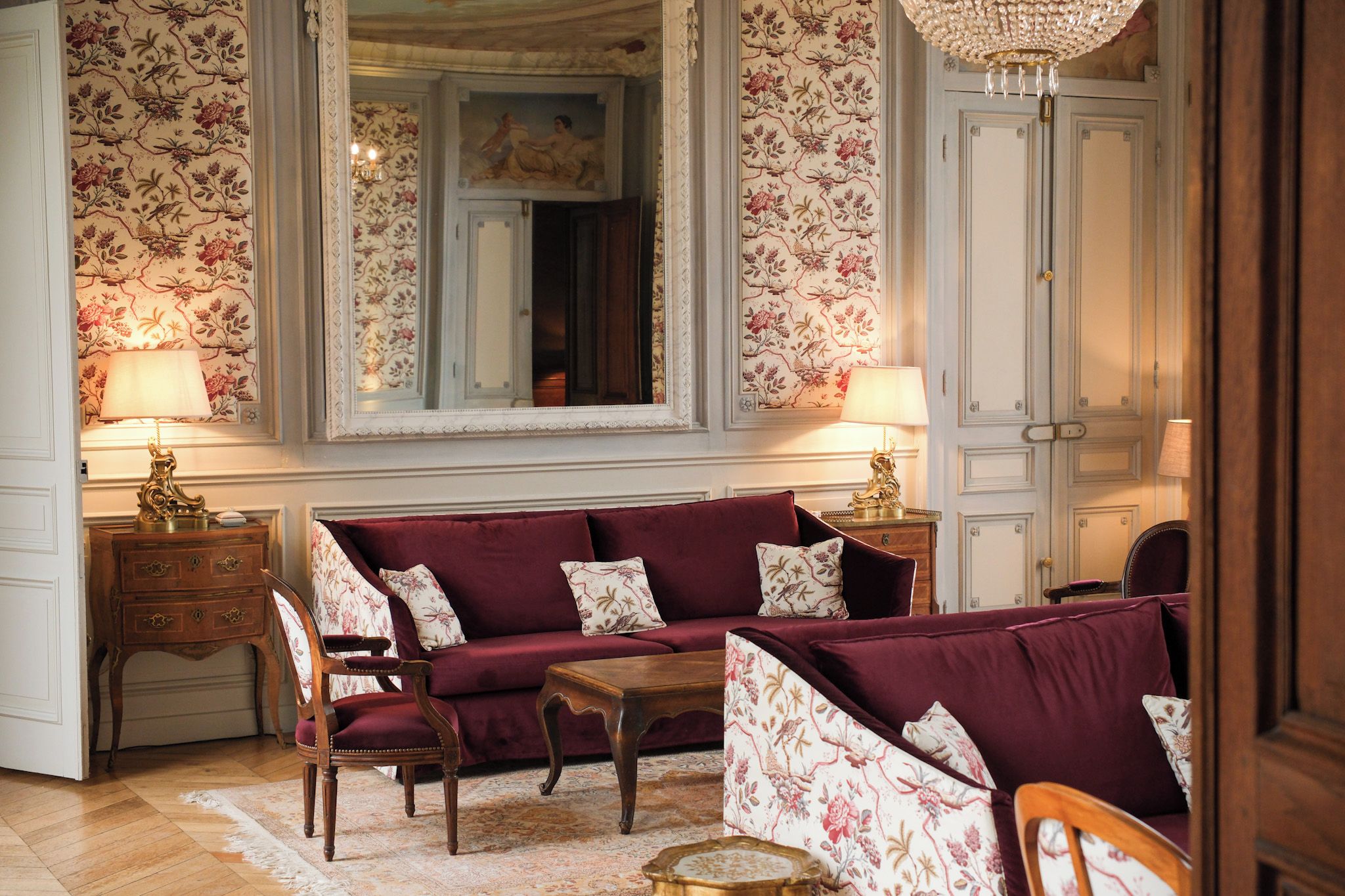 A room with a red sofa, matching armchair, and a wooden side table with a lamp. A large mirror reflects the room, and a chandelier hangs from the ceiling.