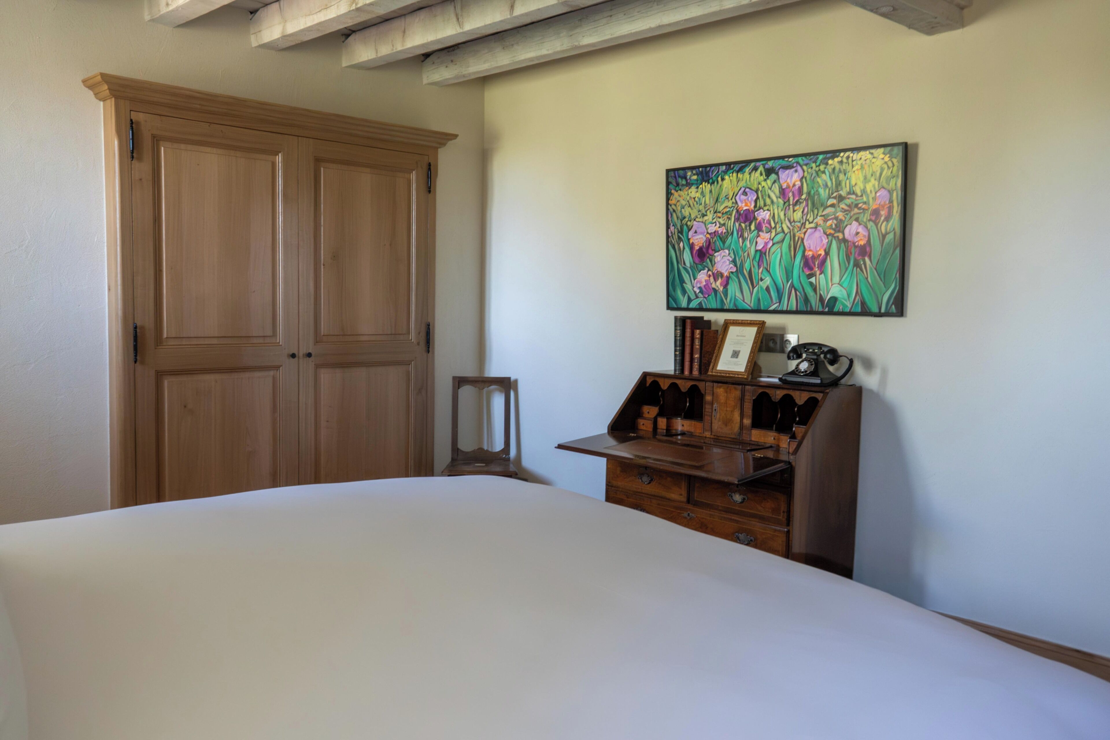 A bedroom with a large bed, wooden wardrobe, a small wooden cabinet with bottles, and a painting of flowers on the wall.