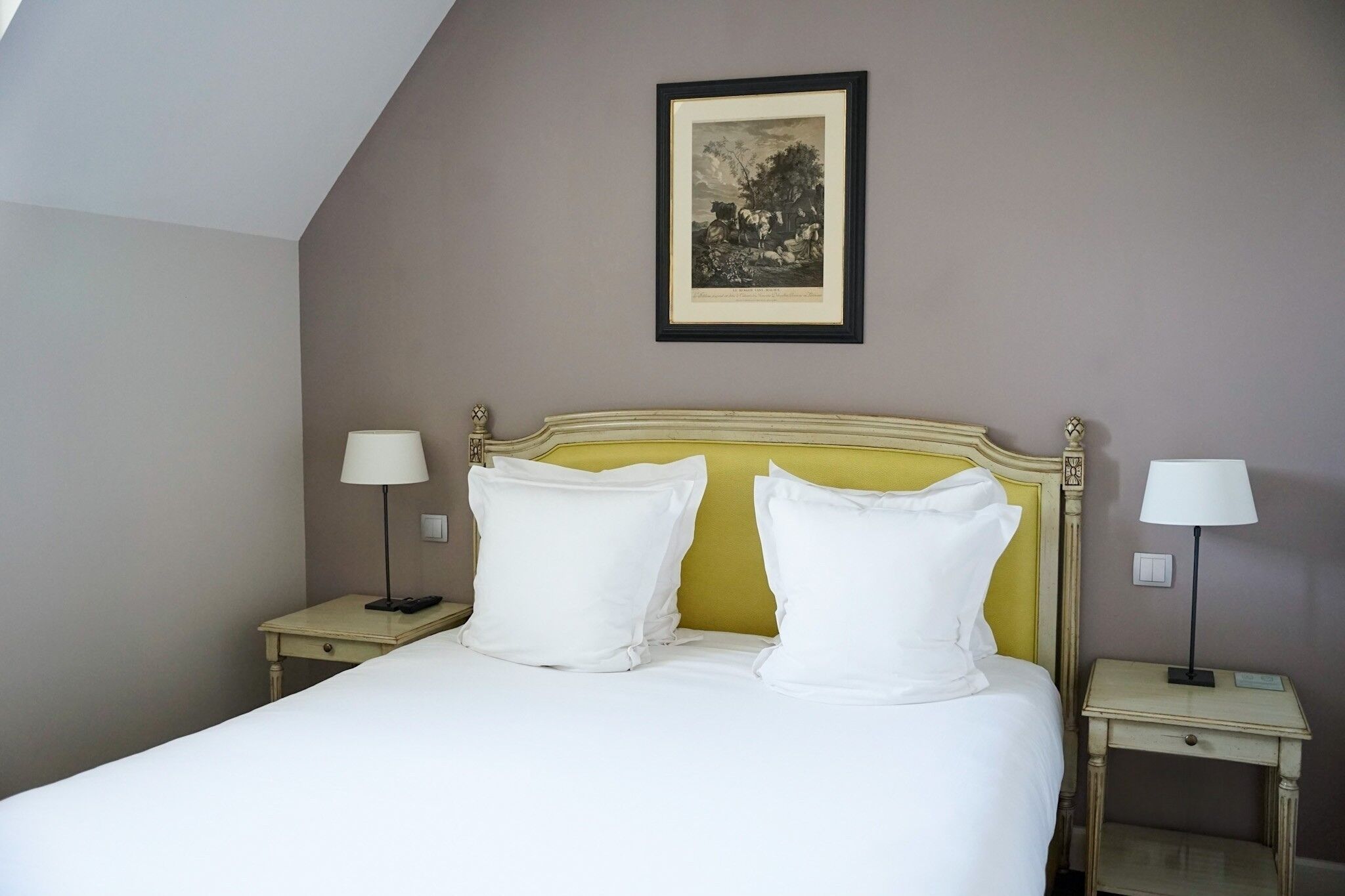 Une chambre d’hôtel avec un lit, deux tables de chevet avec des lampes, un tableau encadré au mur et un plafond en pente.